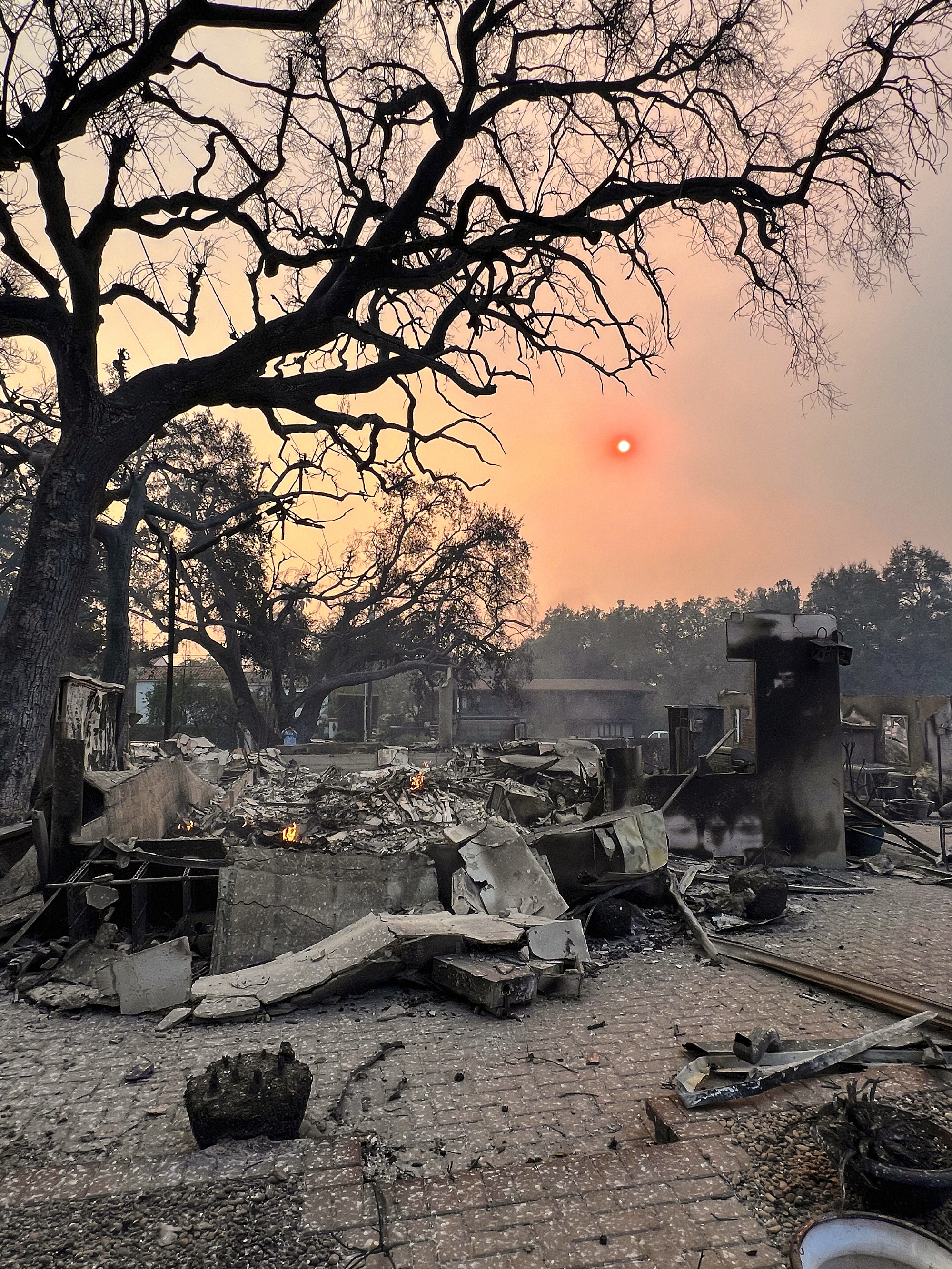 A photo of charred remains of a home with a smoky sky and an orange sun behind them.