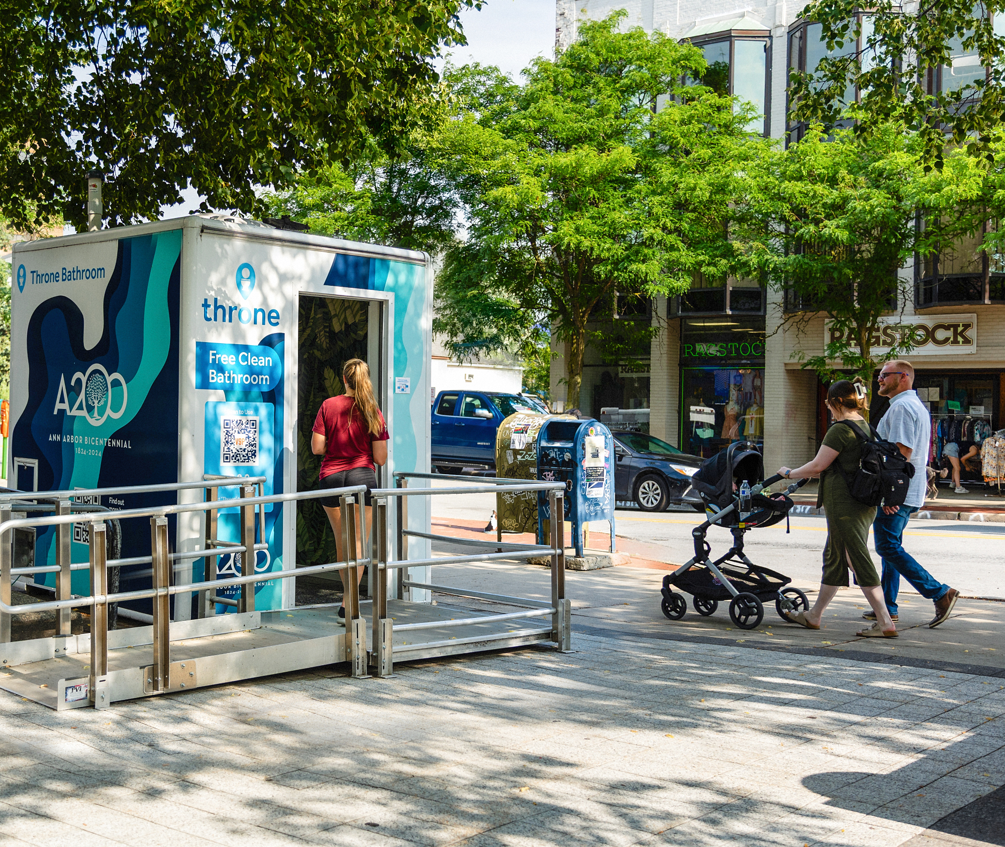 A Throne restroom unit with a person entering and a couple on the street walking by