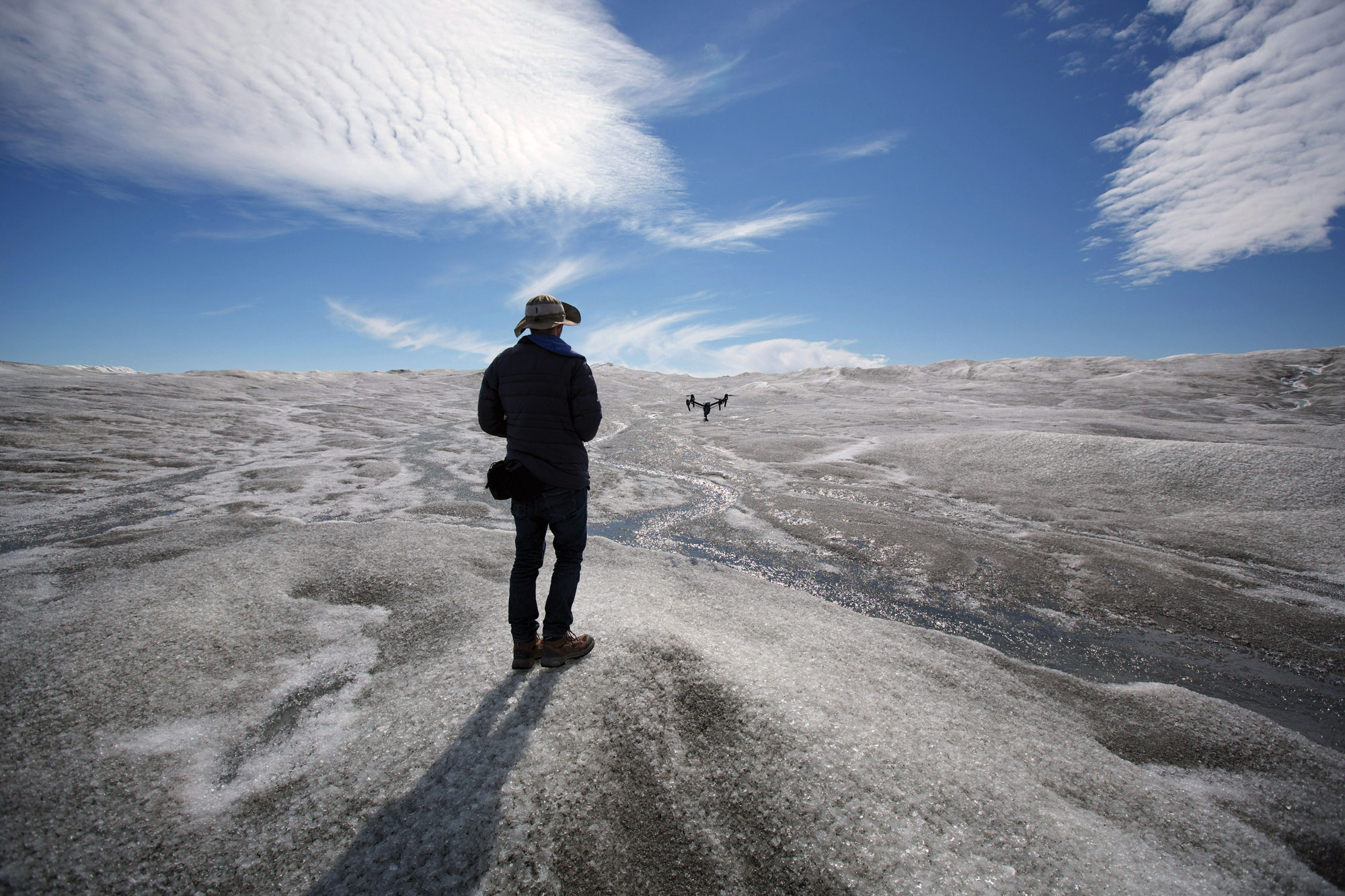 Josh Haner stands on a vast glacier beneath a bright blue sky, operating a drone above meltwater channels carved into the ice.