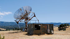 A small shack out on a hill. Standing next to it is a large satellite dish pointed outwards.