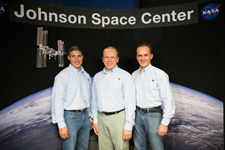 Hopkins, Kotov, and Ryazanskiy stand together in front of a large picture of the Earth and a space shuttle. The picture is labeled 'Johnson Space Center.'