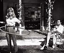 A man in a white suit is seen holding what appears to be a book. In front of him is a young woman in pants and a button down twirling a hula hoop.