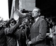 Richard Lyman is speaking through a megaphone a younger man is holding up to his face. He is surrounded by a crowd of people, some of whom hold cameras.