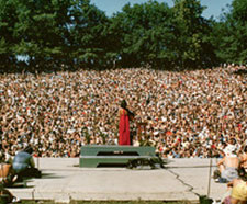 The Grateful Dead stand on the stage of Frost Amphitheater on a bright, sunny day. The crowd is very densely packed, covering the entire lawn.