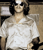 A guard from the Stanford prison experiment. He wears dark aviators and a silky button-down shirt.