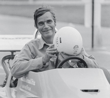 Hargadon is seated in a race car with a small, raised cockpit. He holds a racing helmet outwards.