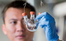 In the foreground is Cui's blue-gloved hand, holding a 3-capped glask flask. Connected to the left and rightmost caps are two wires.