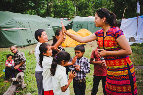A female volunteer worker high fives a boy over a group of young children. They are all well-dressed or in what appear to be school uniforms. To the left, a man sits on a log with a younger girl in his lap. Behind them are a series of tents.