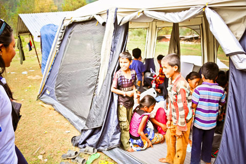 Children are gathered inside a large-tent. Many are sitting in a circle, while others are at the door, looking to an adult volunteer.