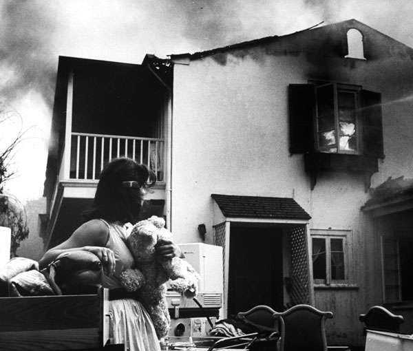 Loftus stands in front of a burning home, clutching a teddy bear tightly to her chest. Furniture and appliances are piled all around the outside.