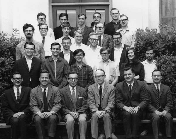 Loftus sits among a throng of well-dressed men. She is in the second row, with sunglasses over her head and her arms folded.