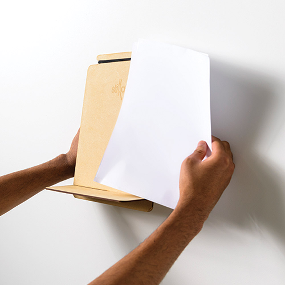 Close-up photo of Dinakar’s invention “Stikato,” a golden-colored music stand about the size of a sheet of paper with a small shelf extending out the bottom. A pair of hands demonstrate its size next to a white sheet of paper.