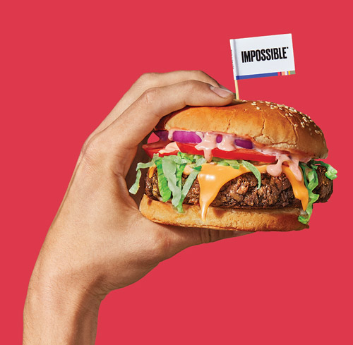 Hand holding an Impossible Burger with a toothpick flag that reads "Impossible".