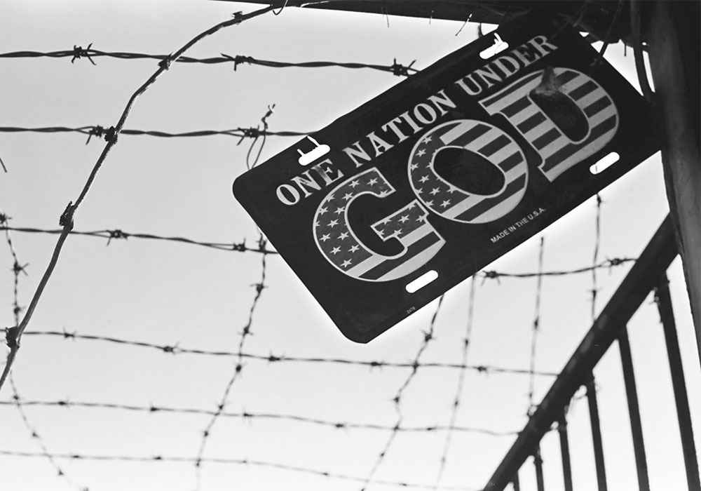 A metal sign that reads, "One Nation Under God", hanging against a barbed wired fence.
