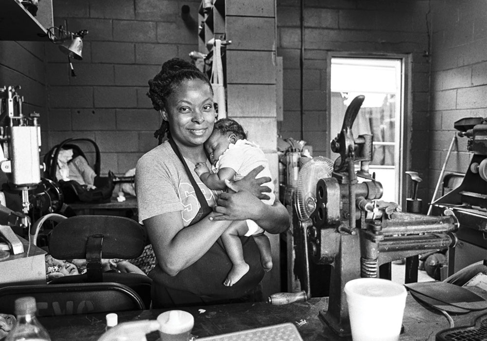 A smiling African Amercian woman in an apron, holding an infant in her work area.