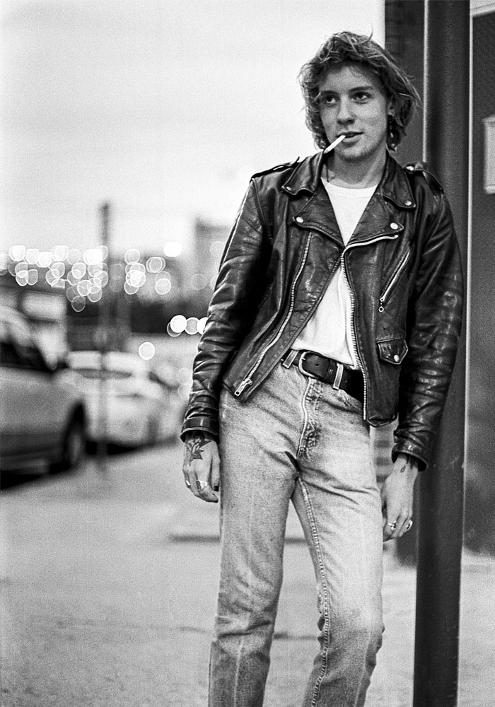 A young, Caucasian man leaning against a pole while smoking and wearing a jean jacket and jeans.