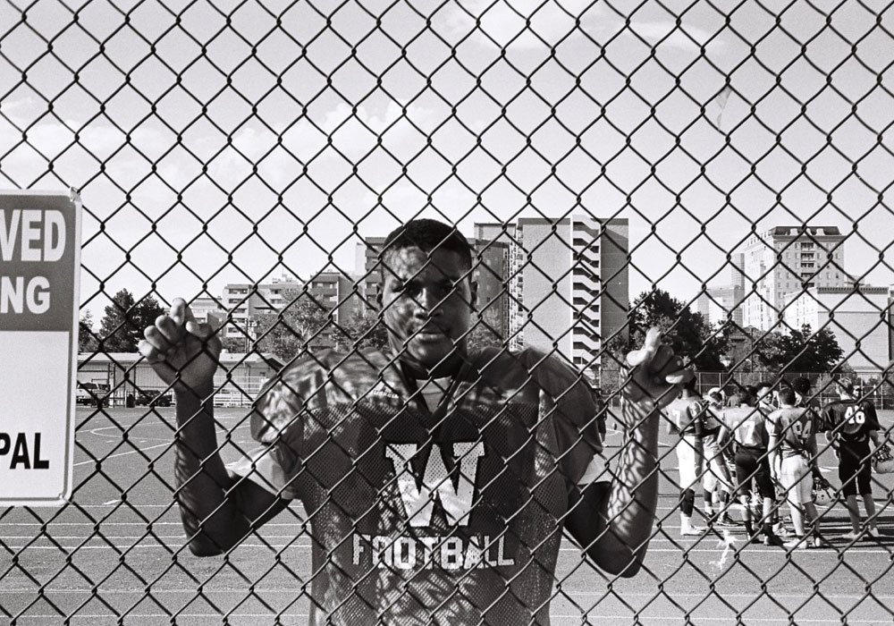 A young, African American male in a football uniform, holding on to a fence on a football practice field.