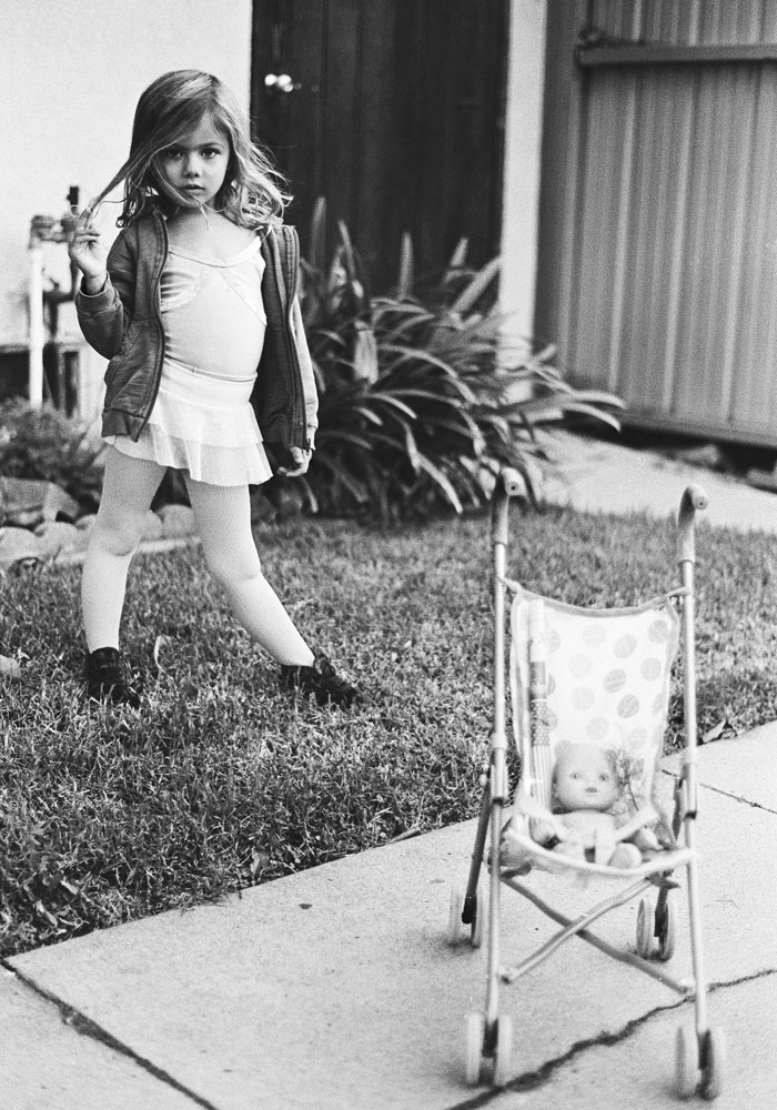 A young girl standing in her front yard, wearing a ballerina outfit and sweatshirt. Her doll and carriage sit on the sidewalk in front of her.