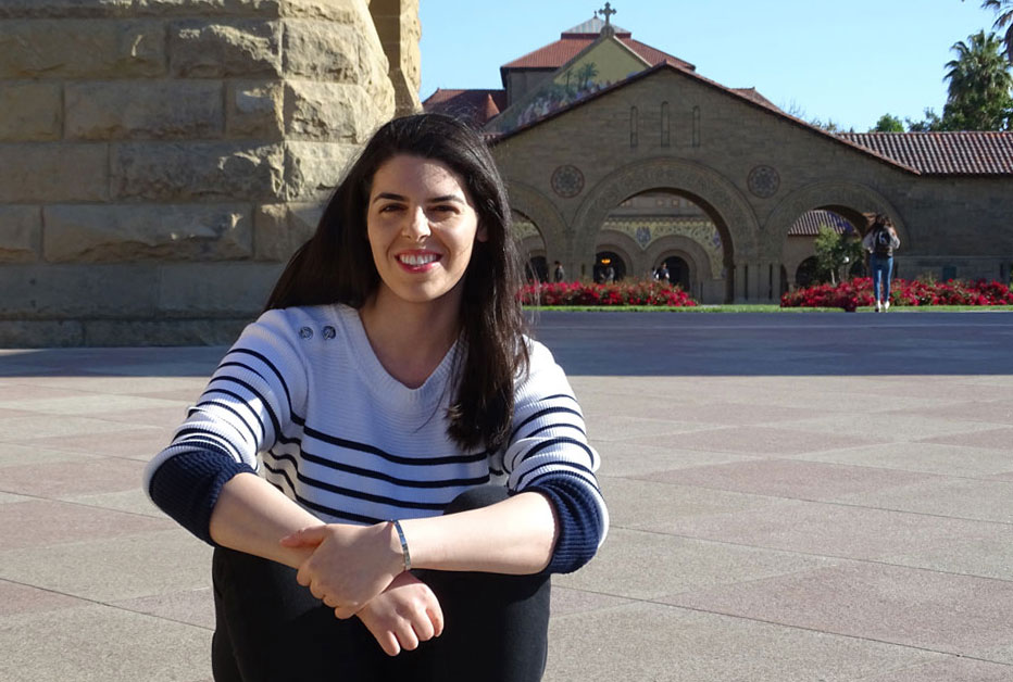 Abdullah smiling and sitting on the steps of the Main Quad of Stanford with Stanford Memorial Church in the background.