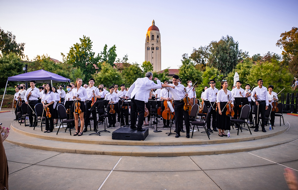The conductor of the Stanford Summer Symphony bumping elbows with with doctoral student Alexander Hwang. The rest of the symphony stands on stage with Hoover Tower in the background.