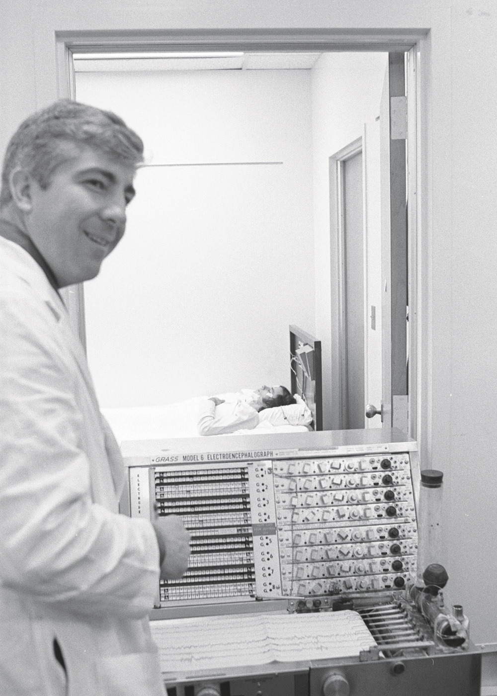 Dement standing by a machine and someone sleeping in the background