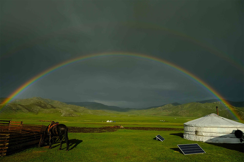 A rainbow arching over green fields; a horse and a yurt with solar panels in the foreground.