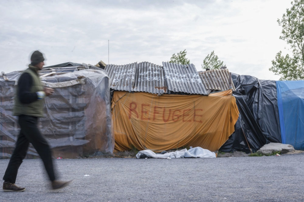 A person walking past a makeshift shelter that says "refugee."