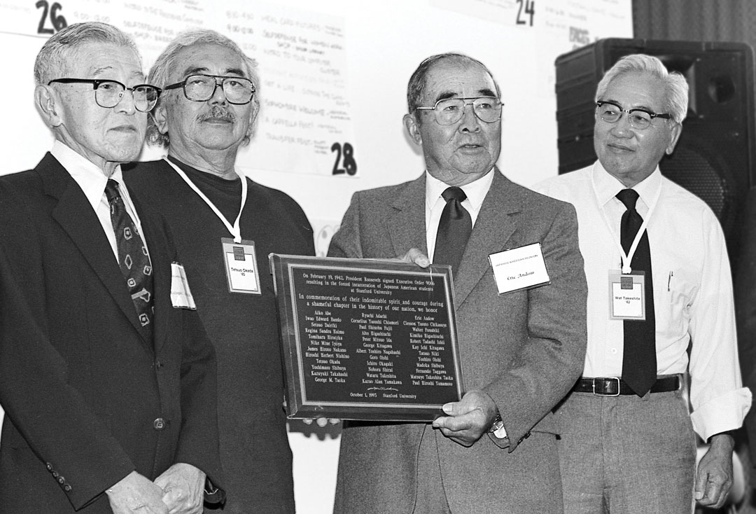 Four Japanese American alumni stand together displaying a commemorative plaque honoring Stanford students affected by World War II internment.