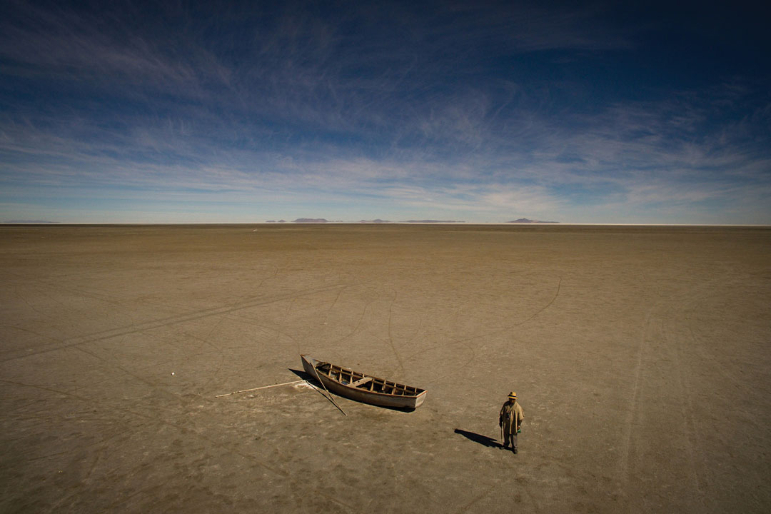 A lone figure stands beside a wooden boat stranded on a vast dry lakebed, with distant mountains stretching across the horizon beneath a sweeping sky.