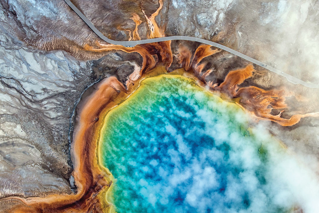 Aerial view of a vivid blue geothermal pool ringed with orange, yellow, and brown mineral deposits, partially veiled by drifting clouds.