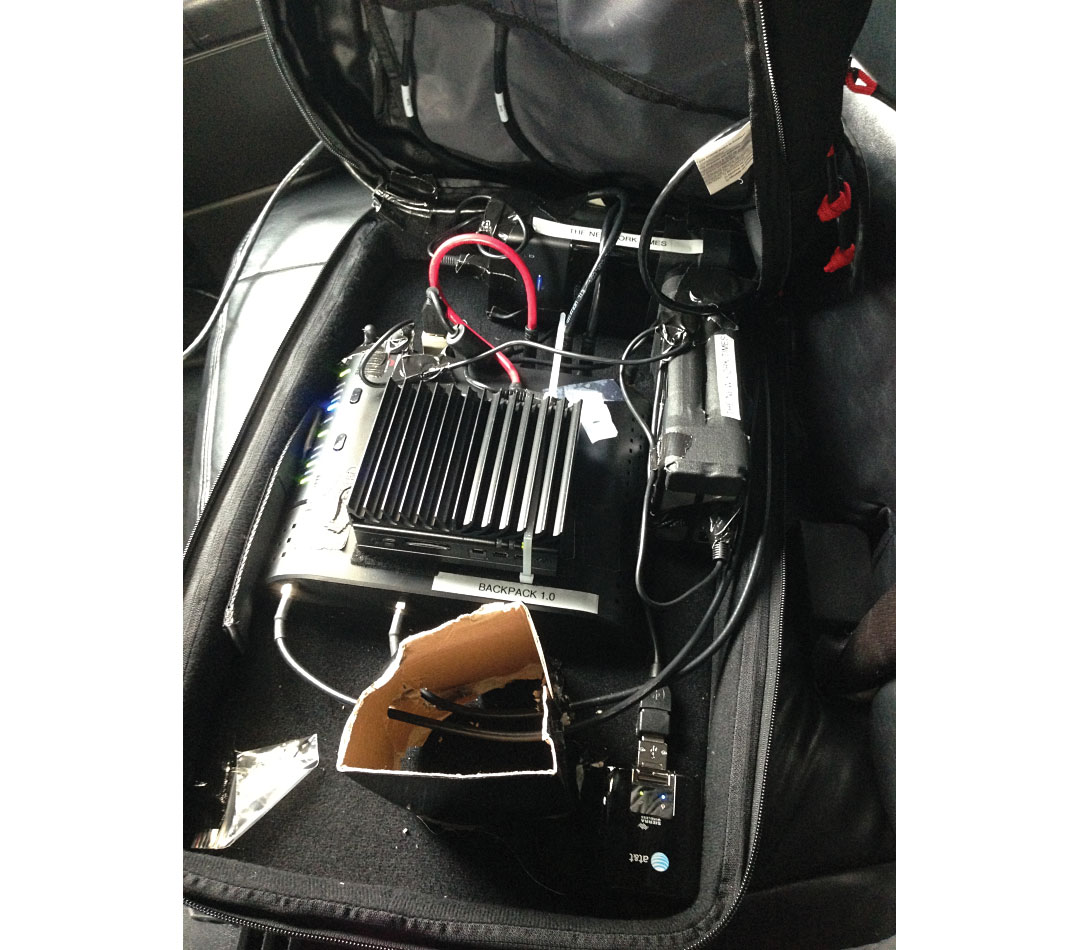 An open backpack contains a portable computing and networking setup, with cables, power components, and electronic equipment packed inside for field reporting.
