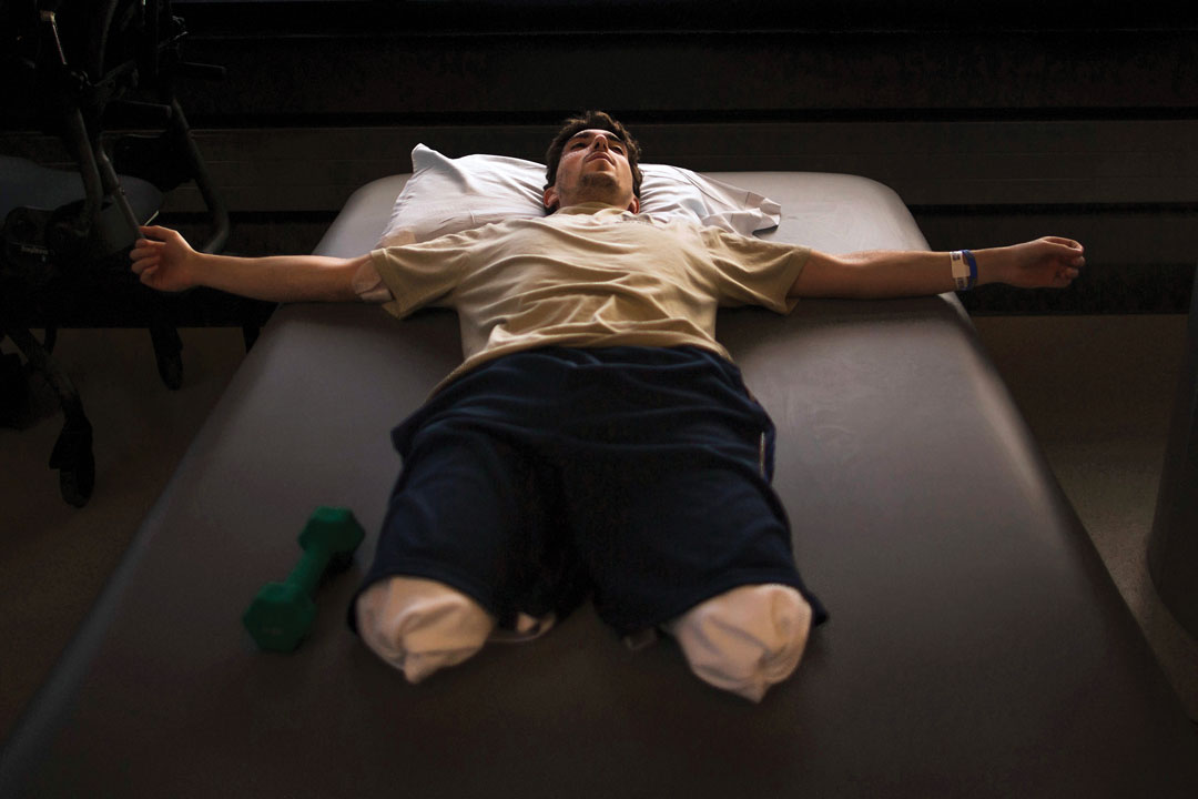 Jeff Bauman lies on a rehabilitation table with his arms outstretched, bilateral leg amputations wrapped in bandages, a hand weight beside him during physical therapy recovery.
