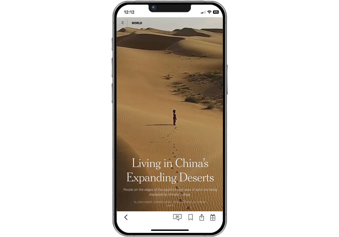 A smartphone displays a feature story titled “Living in China’s Expanding Deserts” over a photograph of a lone figure standing among sweeping sand dunes.