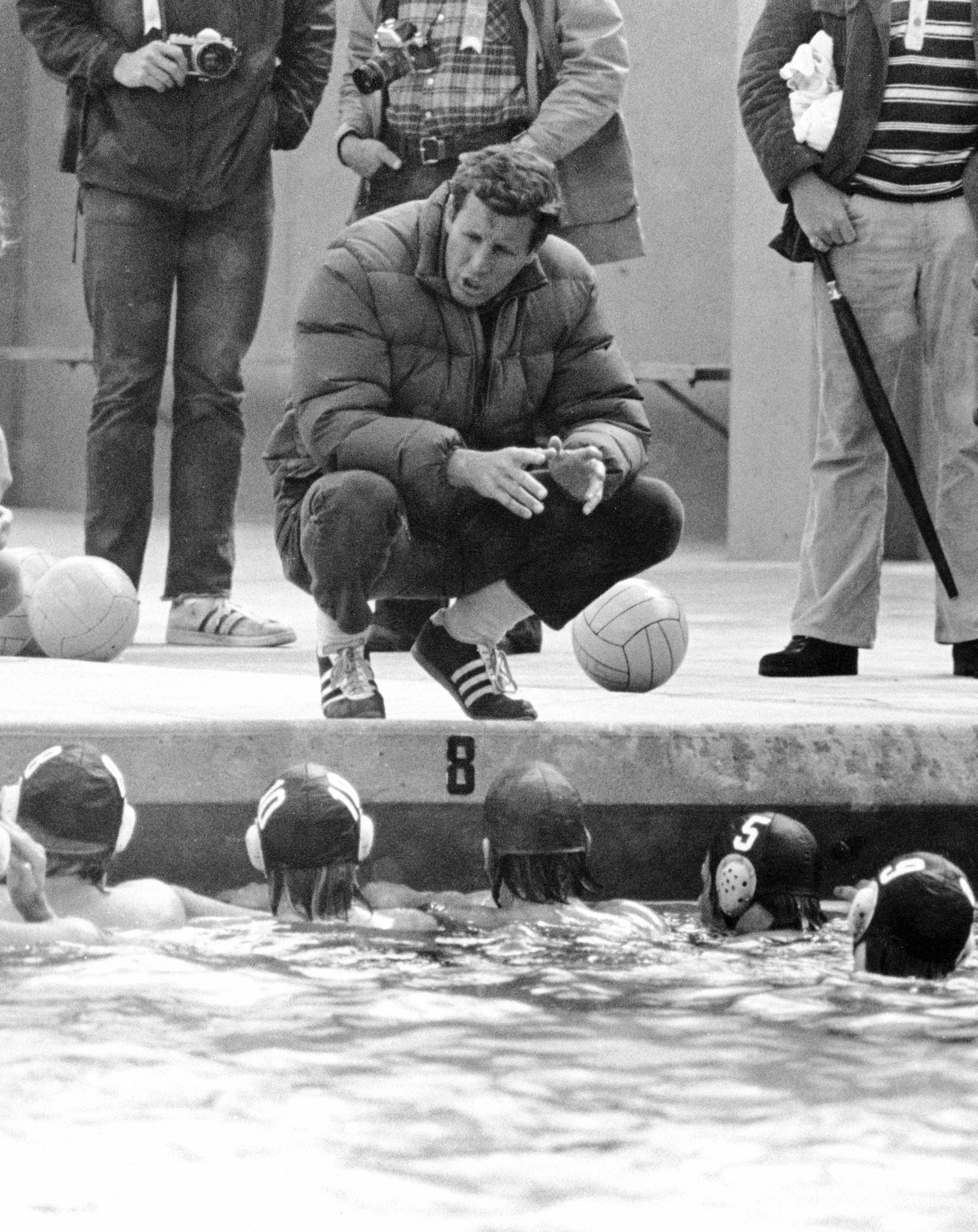 Lambert crouching next to swimming pool, giving directions to the men's water polo team.