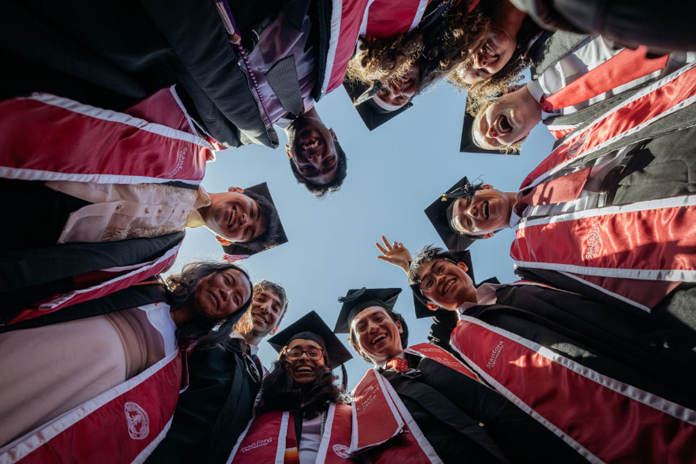 A group of graduates at Commencement in a circle looking down to the camera