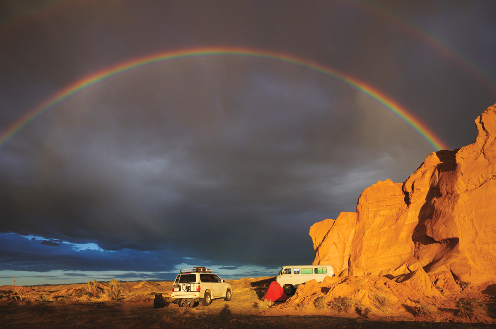 Two vehicles parked by a rock formation with a rainbow in the background