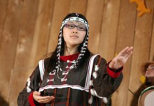 A woman in a feathered headdress wearing black, red, and white garb holds one arm out and one close to her stomach.